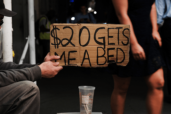 Man holding cardboard sign saying "$20 get me a bed"